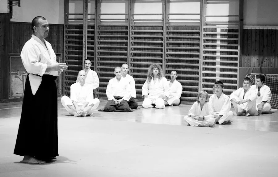 Tréner v hakame vysvetľuje techniku, dospelí aj deti v kimonách sedia na tatami a počúvajú